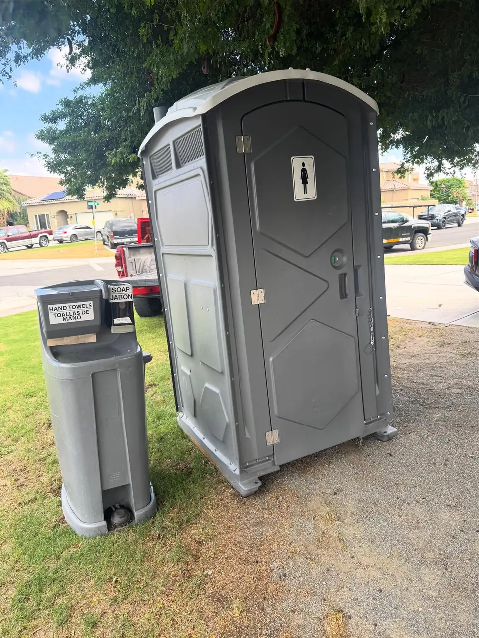 Gray porta potty with a two-sink hand wash station on a lawn